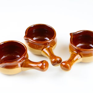 May include: Three brown and beige ceramic soup bowls with handles. The bowls are glazed and have a rustic look.