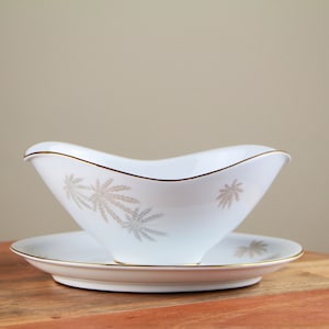 May include: A white porcelain gravy boat with a gold trim and a delicate floral pattern. The boat is sitting on a matching saucer.