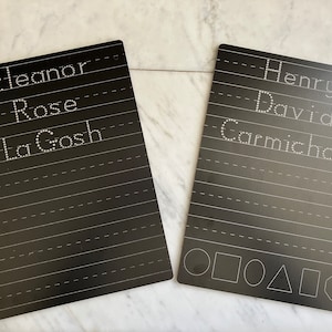 May include: Two black dry-erase boards with white dotted lines for writing practice. One board has the names "Eleanor", "Rose", and "LaGosh" written on it. The other board has the names "Henry", "David", and "Carmichael" written on it. Both boards have geometric shapes at the bottom.