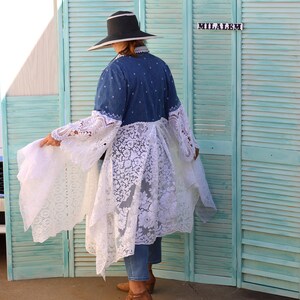 May include: A woman wearing a denim jacket with a white lace overlay and a wide-brimmed black hat. She is standing in front of a teal blue shuttered screen with the word "MILALEM" written on it.