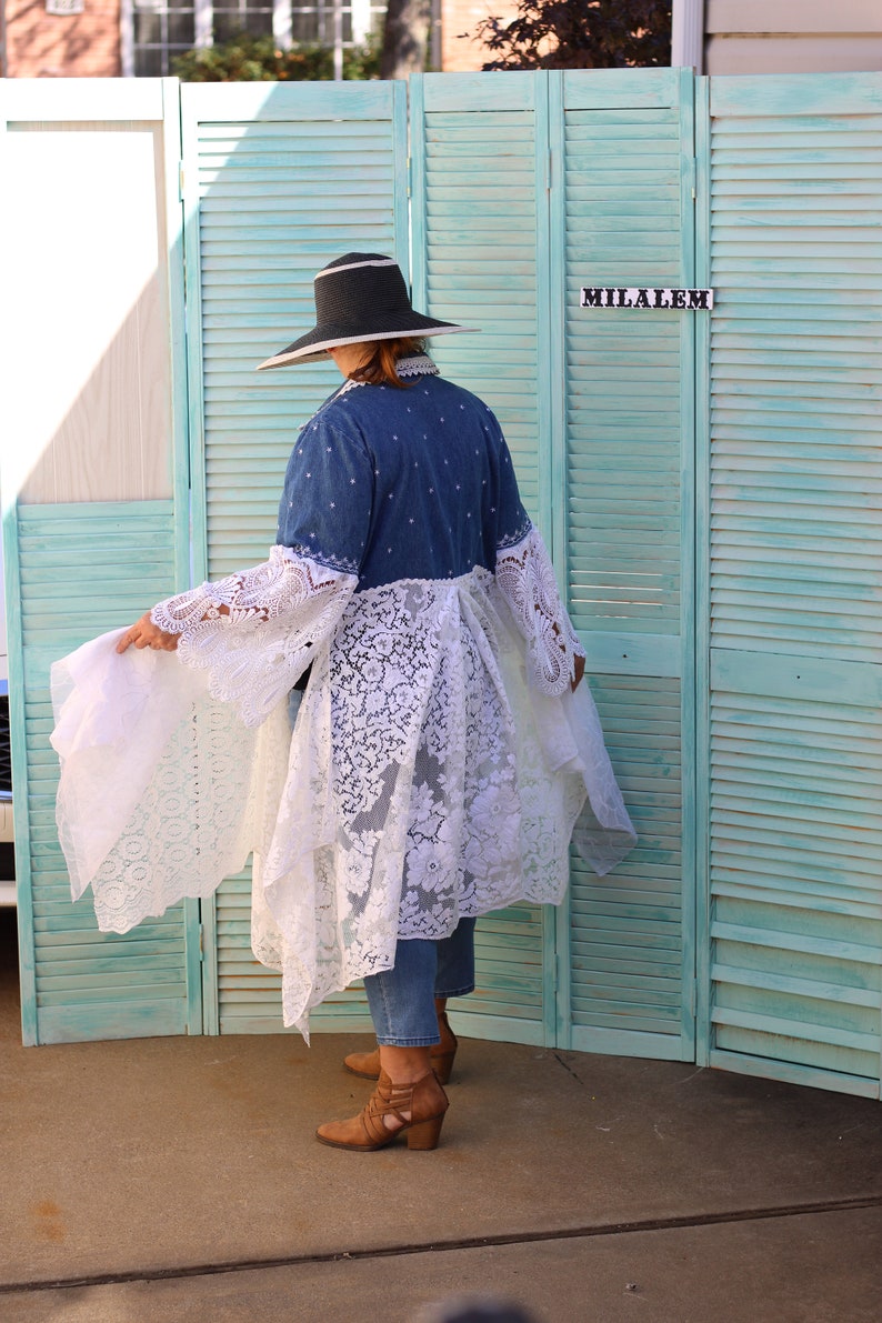 May include: A woman wearing a denim jacket with a white lace overlay and a wide-brimmed black hat. She is standing in front of a teal blue shuttered screen with the word "MILALEM" written on it.