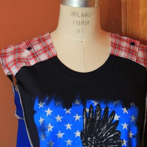 May include: A black sleeveless top featuring red and white plaid fabric panels on the shoulders with dark buttons. The front displays a blue rectangular graphic with white stars and a black wing illustration, worn on a beige dress form.