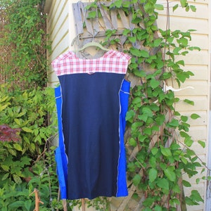 May include: A sleeveless dress featuring a solid navy blue center panel, bright blue side panels, and a red and white plaid yoke at the neckline. The garment is constructed with visible exposed seams along the sides and hangs from a hanger.