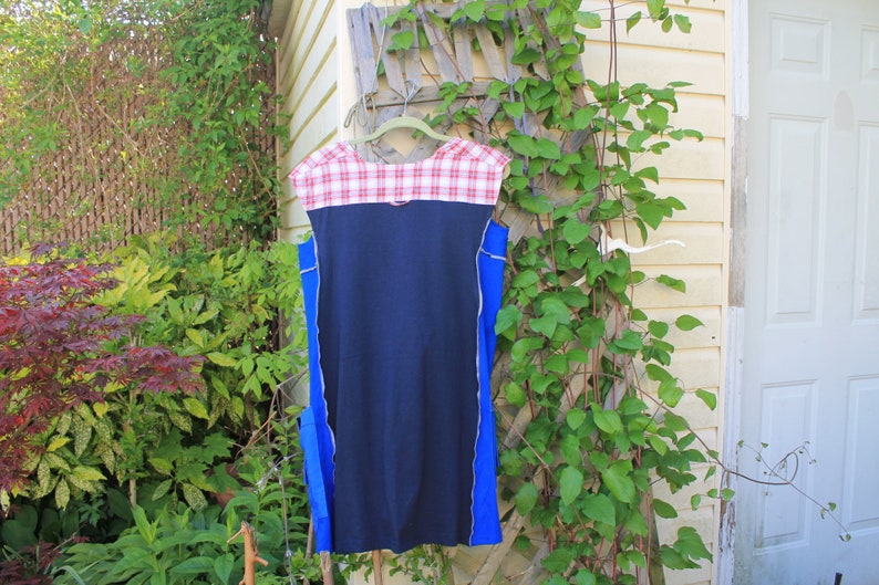May include: A sleeveless dress featuring a solid navy blue center panel, bright blue side panels, and a red and white plaid yoke at the neckline. The garment is constructed with visible exposed seams along the sides and hangs from a hanger.