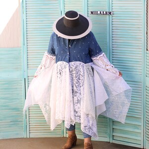 May include: A woman wearing a denim shirt with a white lace overlay and a wide-brimmed black hat. She is standing in front of a teal blue wooden screen with the word "MILALEM" written on it.