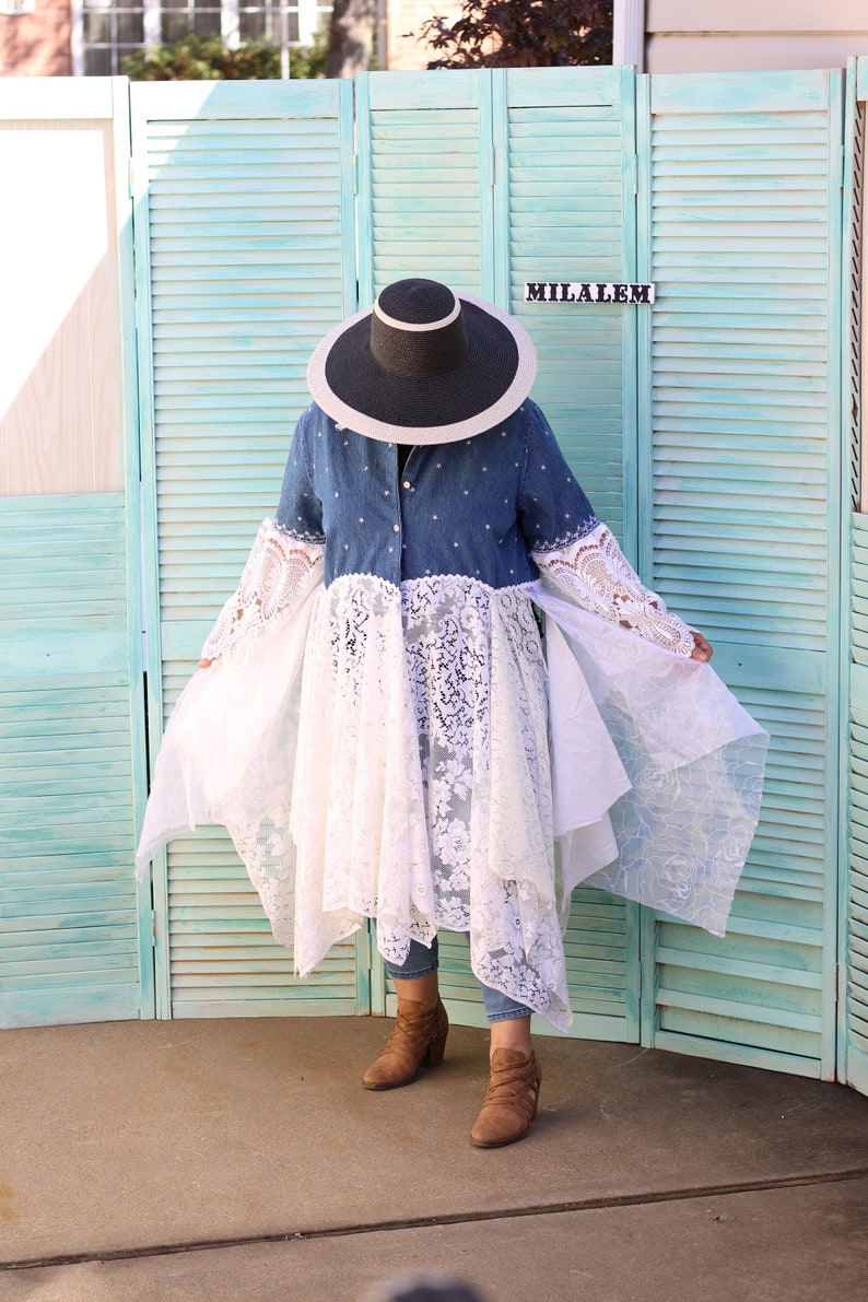 May include: A woman wearing a denim shirt with a white lace overlay and a wide-brimmed black hat. She is standing in front of a teal blue wooden screen with the word "MILALEM" written on it.