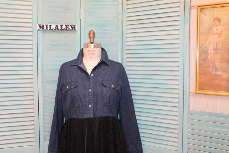 May include: A denim shirt dress with a black lace skirt overlay. The dress is on a mannequin in front of a light blue slatted wall.