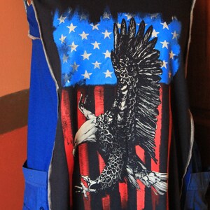 May include: A black sleeveless garment featuring a large graphic print of a bald eagle in flight against a stylized American flag background with blue stars and red and white stripes. The piece includes blue side panels and visible stitched seams.