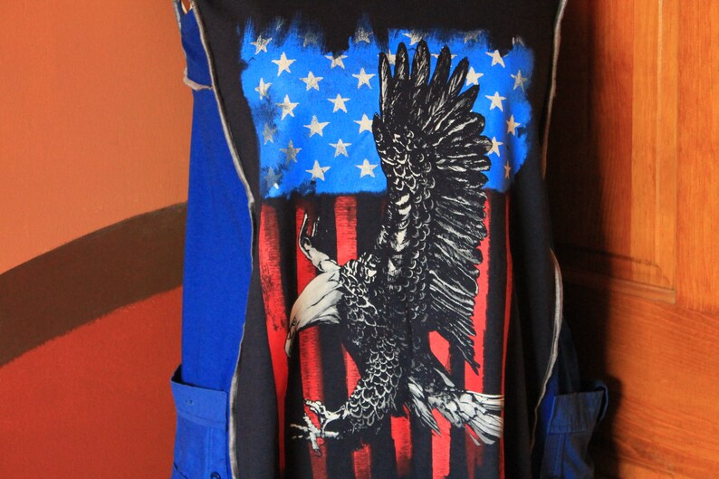 May include: A black sleeveless garment featuring a large graphic print of a bald eagle in flight against a stylized American flag background with blue stars and red and white stripes. The piece includes blue side panels and visible stitched seams.