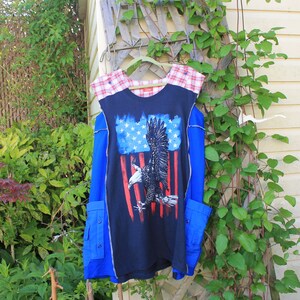 May include: A sleeveless navy blue tunic featuring a central graphic of a bald eagle superimposed over a stylized American flag. The garment includes red and white plaid shoulder panels, blue side panels, and two lower pockets with button closures.