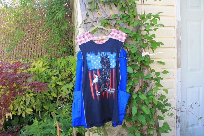 May include: A sleeveless navy blue tunic featuring a central graphic of a bald eagle superimposed over a stylized American flag. The garment includes red and white plaid shoulder panels, blue side panels, and two lower pockets with button closures.