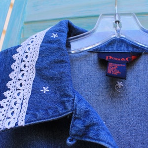 May include: A blue denim shirt with a white lace collar and embroidered white stars. The shirt has a label that reads "Denim & Co."