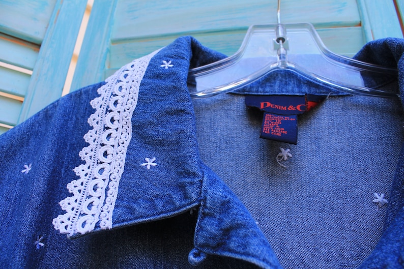 May include: A blue denim shirt with a white lace collar and embroidered white stars. The shirt has a label that reads "Denim & Co."