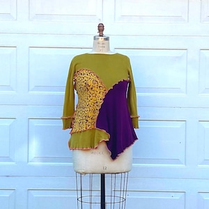 Könnte beinhalten: Olivgrünes Langarm-Oberteil mit einem einzigartigen asymmetrischen Design. Das Oberteil hat einen Abschnitt aus gelbem Blumenstoff und eine tiefviolette Partie. Die Kanten sind mit einer kontrastierenden orangefarbenen Naht versehen, was ein optisch auffälliges Kleidungsstück ergibt.