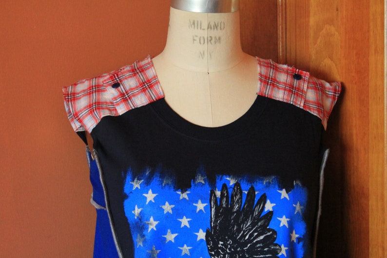 May include: A black sleeveless top featuring red and white plaid fabric panels on the shoulders with dark buttons. The front displays a blue rectangular graphic with white stars and a black wing illustration, layered over a solid blue base.