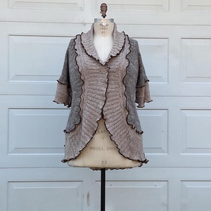 Op de afbeelding: Gebreid vest in tinten grijs en beige, weergegeven op een mannequin. De open trui heeft een gerimpelde kraag en bies in donkerder bruin, met korte mouwen en een enkele knoopsluiting. Het kledingstuk is maat 22.