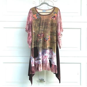 May include: A tunic top with a scoop neckline and short, flared sleeves. The garment features a brown and pink tie-dye pattern with chocolate-themed graphics and text including "CHOCOLATE" and "AMERICA'S FAVORITE". It has a dark brown pocket.