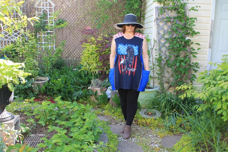 May include: A sleeveless navy blue tunic featuring a graphic of a bald eagle in front of red and white stripes, red and white plaid shoulder panels, and blue side pockets, paired with a wide-brimmed hat, sunglasses, and black leggings.