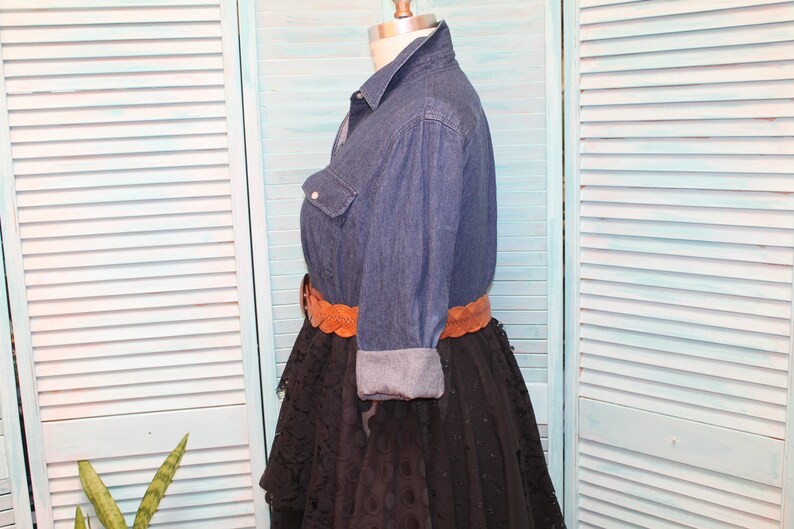 May include: A denim shirt with long sleeves and a brown leather belt. The shirt is worn over a black lace skirt.