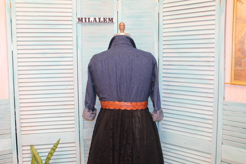 May include: A blue denim shirt with long sleeves and a brown braided belt. The shirt is on a mannequin in front of a light blue wooden slatted background.