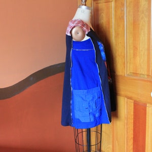 May include: A sleeveless tunic top featuring a color-blocked design with black and royal blue panels. The garment includes a red and white plaid shoulder detail and a large blue utility pocket on the side. It is displayed on a dress form mannequin.