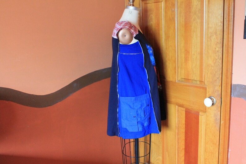 May include: A sleeveless tunic top featuring a color-blocked design with black and royal blue panels. The garment includes a red and white plaid shoulder detail and a large blue utility pocket on the side. It is displayed on a dress form mannequin.