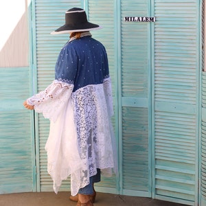 May include: A person wearing a wide-brimmed black hat, a denim jacket with lace trim, and brown boots. The person is standing in front of a teal blue wooden screen with the word "MILALEM" written on it.