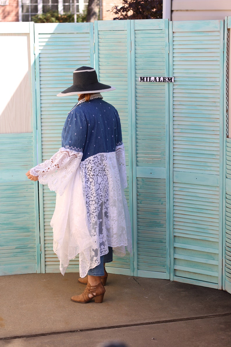 May include: A person wearing a wide-brimmed black hat, a denim jacket with lace trim, and brown boots. The person is standing in front of a teal blue wooden screen with the word "MILALEM" written on it.