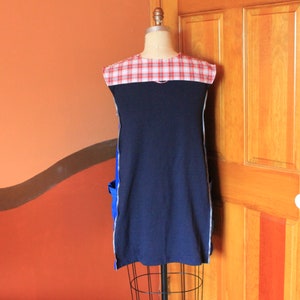 May include: A sleeveless tunic featuring a solid navy blue main body with a red and white plaid yoke at the shoulders. The garment includes bright blue side pockets and white vertical trim along the sides, displayed on a dress form mannequin.