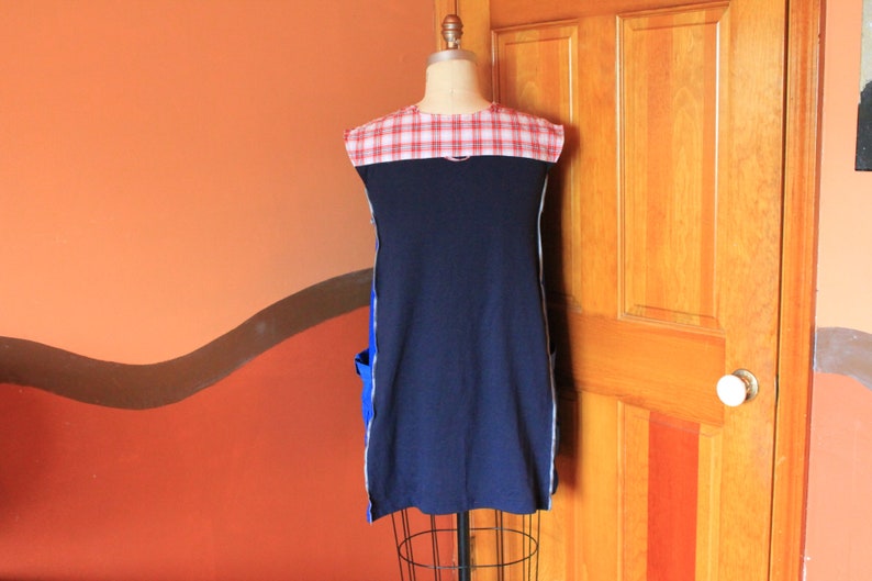 May include: A sleeveless tunic featuring a solid navy blue main body with a red and white plaid yoke at the shoulders. The garment includes bright blue side pockets and white vertical trim along the sides, displayed on a dress form mannequin.