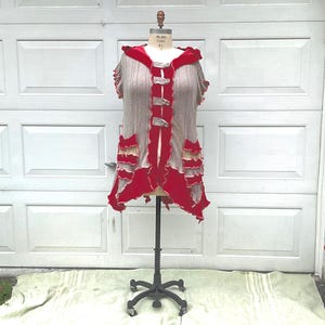 May include: A sleeveless, hooded vest displayed on a dress form. The vest features a gray knit body with red accents, including the hood, trim, and decorative ruffles. It has three button closures down the front.
