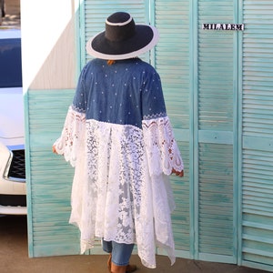 May include: A woman wearing a denim jacket with a white lace overlay and a wide-brimmed black hat. The jacket has a white embroidered design. She is standing in front of a teal blue wooden shutter.
