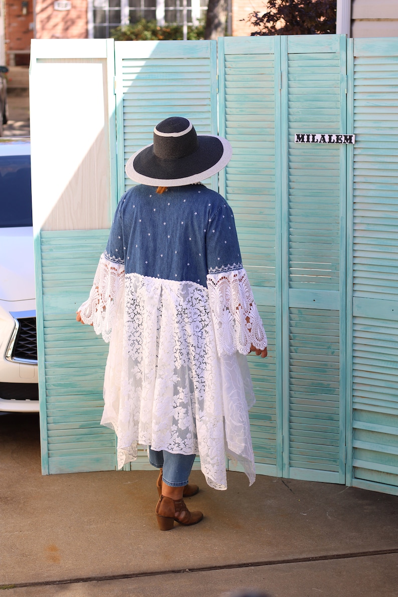 May include: A woman wearing a denim jacket with a white lace overlay and a wide-brimmed black hat. The jacket has a white embroidered design. She is standing in front of a teal blue wooden shutter.