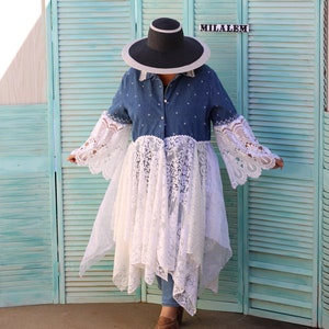 May include: A woman wearing a denim shirt with a white lace overlay, a wide-brimmed black hat, and brown ankle boots. The shirt has a white polka dot pattern. The woman is standing in front of a teal blue wooden slatted screen with the word "MILALEM" written on it.