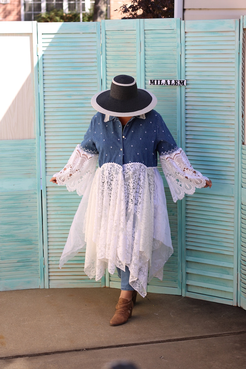 May include: A woman wearing a denim shirt with a white lace overlay, a wide-brimmed black hat, and brown ankle boots. The shirt has a white polka dot pattern. The woman is standing in front of a teal blue wooden slatted screen with the word "MILALEM" written on it.