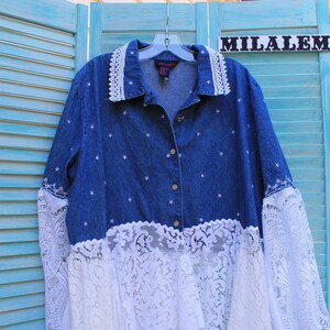 May include: A blue denim shirt with white star embroidery and white lace trim around the collar, cuffs, and bottom hem. The shirt is hanging on a hanger with the word "MILALEM" on a sign above it.
