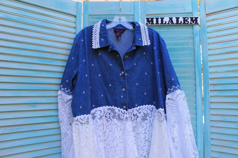 May include: A blue denim shirt with white star embroidery and white lace trim around the collar, cuffs, and bottom hem. The shirt is hanging on a hanger with the word "MILALEM" on a sign above it.