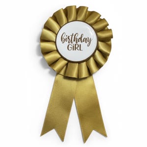 May include: A gold-colored birthday girl rosette with a white center. The center features the words "birthday GIRL" in a brown script font. The rosette has a pleated design and two gold ribbons.