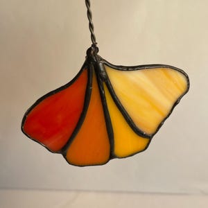 May include: A stained glass ginkgo leaf ornament featuring four distinct segments in a gradient of red, orange, yellow, and cream. The glass pieces are joined by dark metal solder lines and suspended from a twisted metal wire hanger at the top.