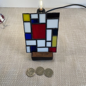 May include: A stained glass lamp in a geometric design with red, yellow, blue, black, and white squares. The lamp sits on a wooden base and has a small light bulb at the top. Three coins are in front of the lamp.