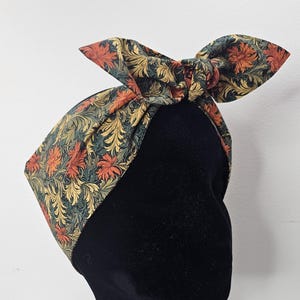 Könnte beinhalten: Ein Stirnband mit floralem Muster und Schleife. Der Stoff zeigt ein Design aus orangefarbenen, grünen und goldenen Blumen und Blättern auf dunklem Hintergrund. Das Stirnband wird auf einem schwarzen Schaufensterkopf präsentiert.