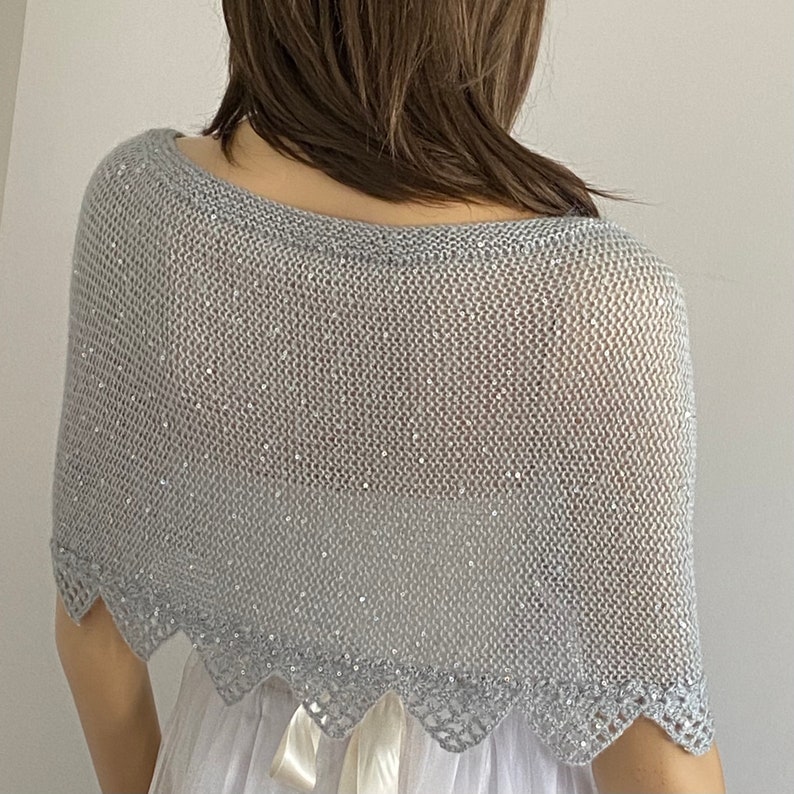 K&ouml;nnte beinhalten: Ein silbergrauer Strickschal mit einem gewellten Rand. Der Schal ist mit kleinen, funkelnden Pailletten verziert, die einen Hauch von Schimmer verleihen. Das offene Webdesign und die filigranen Details machen ihn zu einem stilvollen Accessoire.