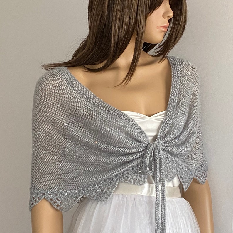 K&ouml;nnte beinhalten: Ein hellgrauer Strickschal mit gewelltem Rand und Bindeband. Der Schal ist mit Pailletten verziert und wird &uuml;ber einem wei&szlig;en Kleid getragen. Der Gesamtstil ist elegant und f&uuml;r besondere Anl&auml;sse geeignet.