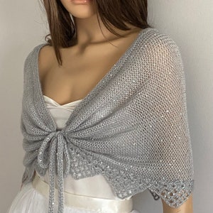 K&ouml;nnte beinhalten: Ein silbergrauer Strickschal mit zartem, offenem Gewebe. Der Schal ist mit Pailletten verziert und hat einen Bindverschluss vorne. Die Kanten haben ein gewelltes Design, das diesem Accessoire einen Hauch von Eleganz verleiht.
