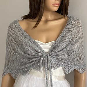 K&ouml;nnte beinhalten: Ein silbergrauer Strickschal mit Bindverschluss. Der Schal hat ein zartes, offenes Webmuster und ist mit Pailletten an den R&auml;ndern verziert. Der Schal ist &uuml;ber einem wei&szlig;en Kleid drapiert und erzeugt einen eleganten Look.