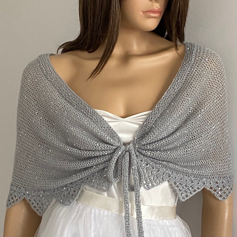 K&ouml;nnte beinhalten: Ein silbergrauer Strickschal mit Bindverschluss. Der Schal hat ein zartes, offenes Webmuster und ist mit Pailletten an den R&auml;ndern verziert. Der Schal ist &uuml;ber einem wei&szlig;en Kleid drapiert und erzeugt einen eleganten Look.