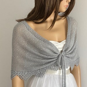 K&ouml;nnte beinhalten: Ein silbergrauer Strickschal mit gewelltem Rand und Bindeband. Der Schal ist mit Pailletten verziert und &uuml;ber einem wei&szlig;en Kleid drapiert. Der Gesamtstil ist elegant und f&uuml;r besondere Anl&auml;sse geeignet.