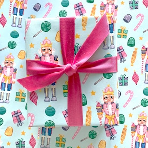 May include: A light blue gift wrap with a pink ribbon. The wrapping paper features a repeating pattern of nutcrackers, candy canes, lollipops, and stars.