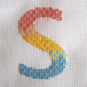 May include: A cross-stitch of the letter 'S' in a rainbow color scheme. The 'S' is made of pink, yellow, and blue thread.