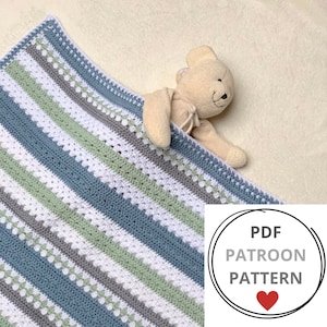 Op de afbeelding: Een gehaakte babydeken met strepen in blauw, groen, wit en grijs. De deken heeft een textuur met kleine witte stippen. De deken is gevouwen en er ligt een knuffeldier op.